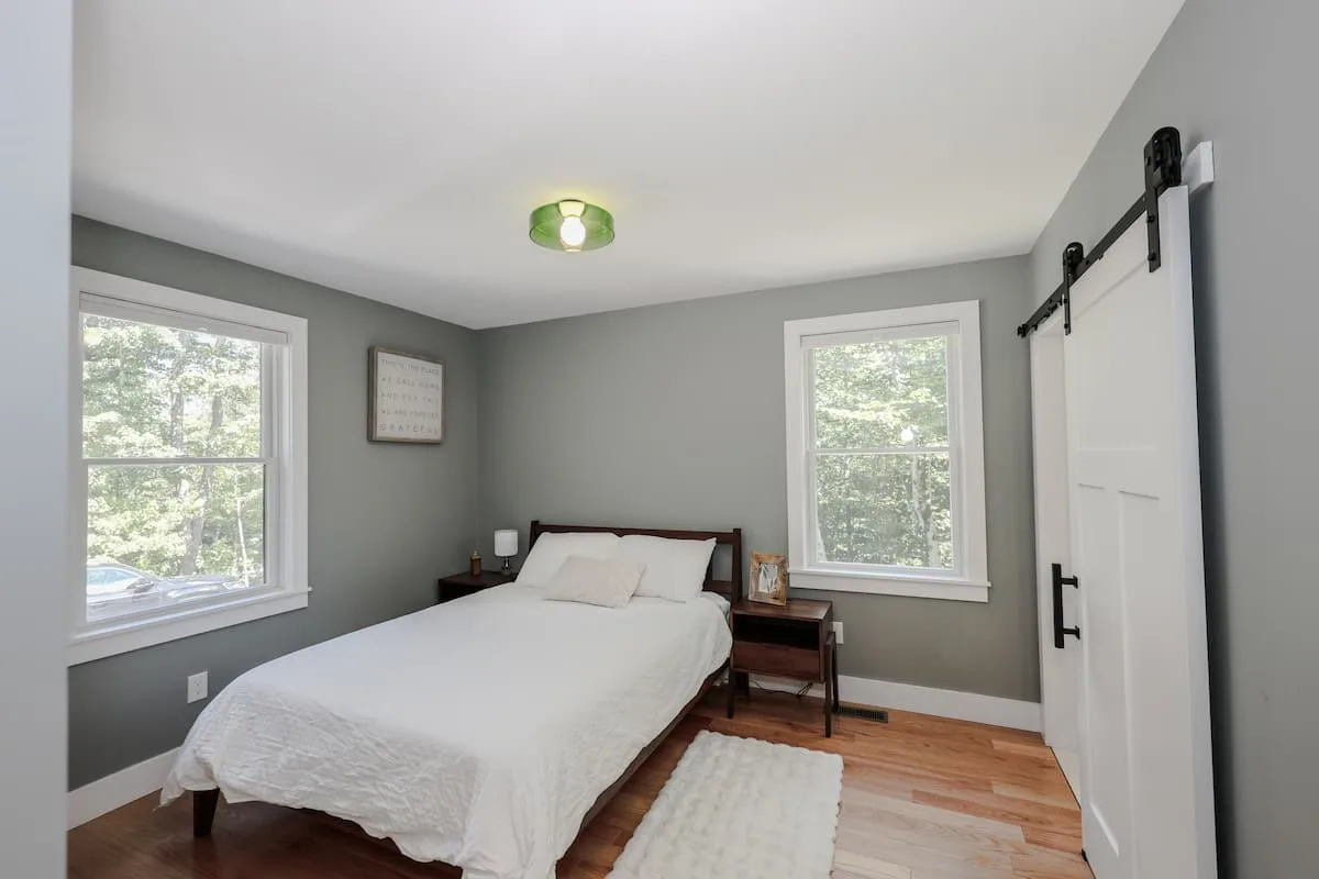 Wood-framed bed with white linens between two large windows beside a white sliding barn door at Grateful Ledge