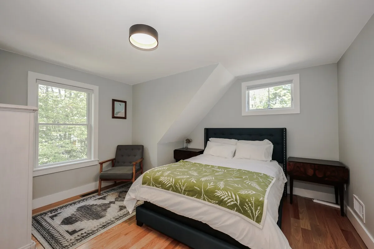 Professionally staged bedroom with a black tufted headboard, green botanical coverlet, two windows, and a sloped wall at Grateful Ledge