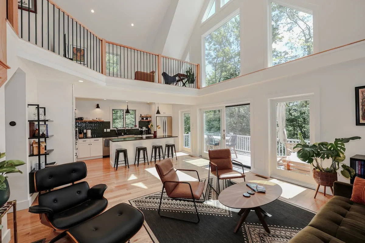 Professionally staged open-plan living room with loft, forest-facing windows, seating, and kitchen beyond at Grateful Ledge