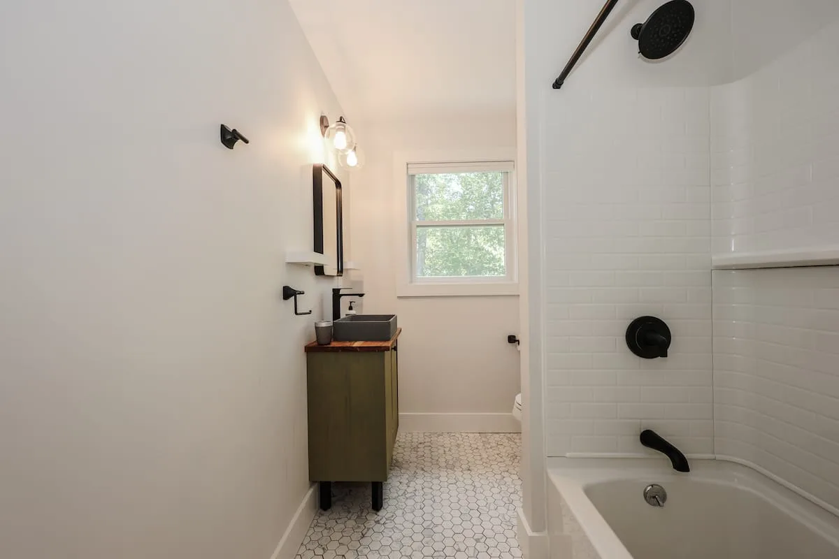 Professionally staged bathroom with a green vanity, white tile tub and shower, hexagonal tile floor, and window at Grateful Ledge